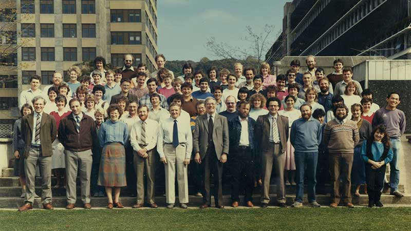 Roughly five rows of people standing on stairs for a group photo with lawn in front of them and two buildings behind them