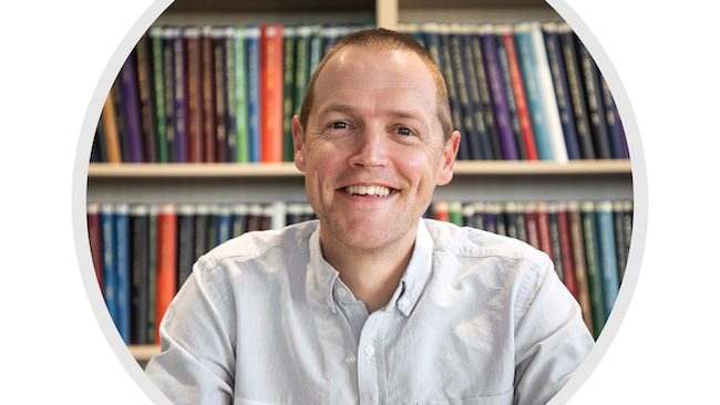 Peter Mace smiling and sitting in front of a bookshelf full of theses.