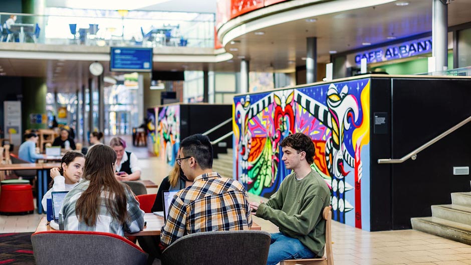 A group of students sitting with laptops at a table in the University of Otago Link area, with Café Albany in the background