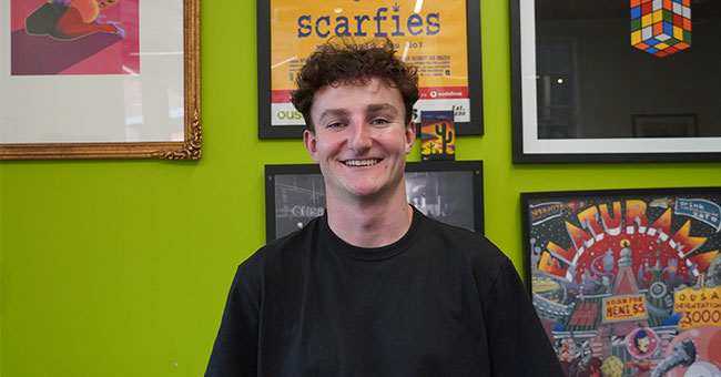 A young man stands in front of a wall of framed OUSA posters.