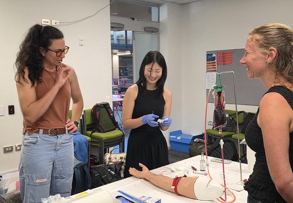 Three students practise inserting an IV cannulation into the arm of a plastic mannequin in a classroom setting.