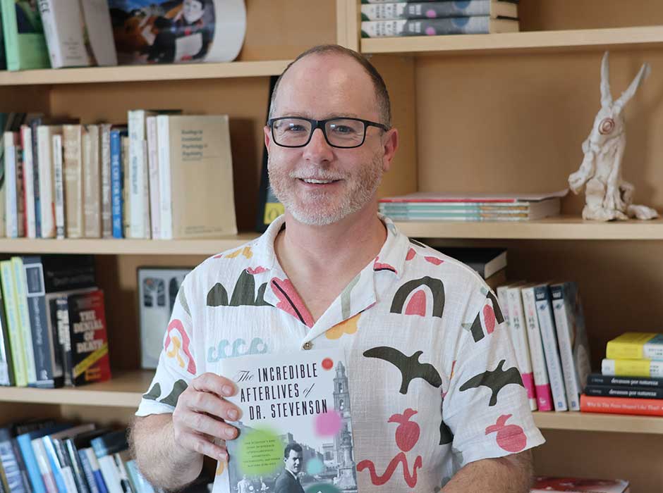 A man from the waist up, holding a book and standing in front of a book case 