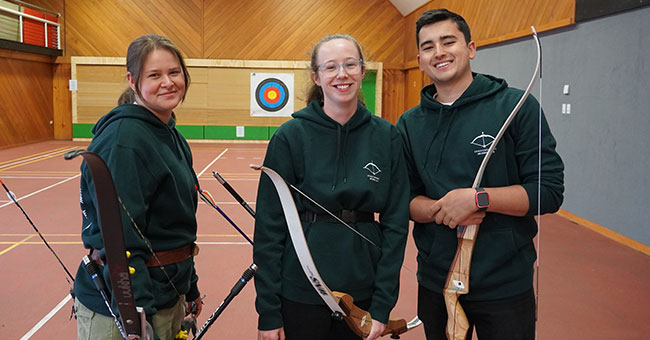 Two women and a man pose with archery equipment in front of an archery wall.