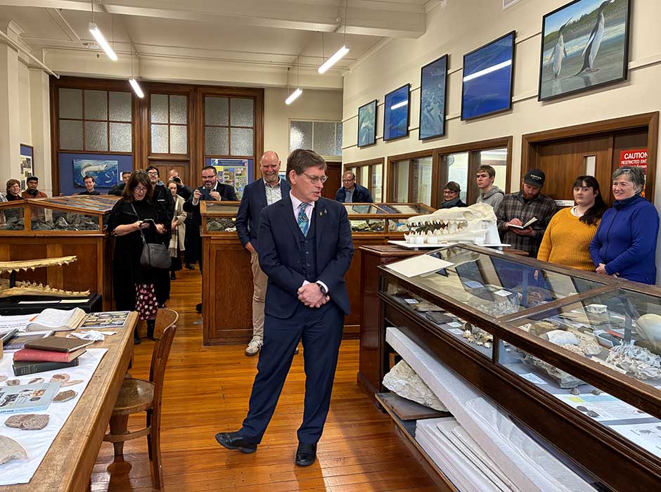 A man standing on a wooden floor in between display cases in a museum speaking to people outside the cases