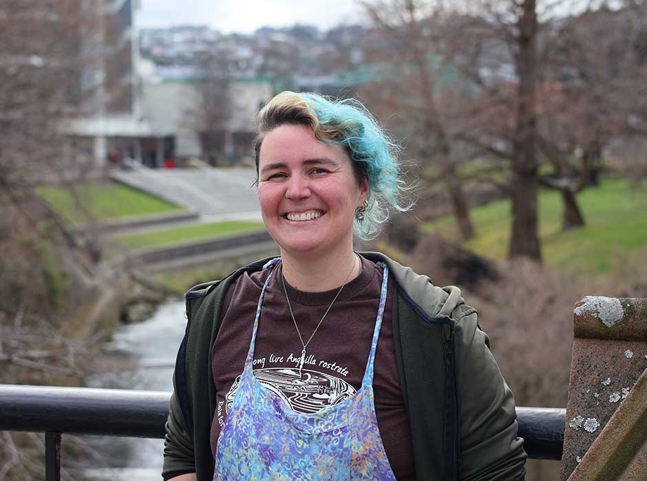 A woman from the waist up standing on a bridge with a river and buildings behind her
