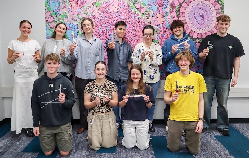 Group photo of eleven smiling students posed in front of a large, colorful scientific illustration mural; most students are standing in the back row while others are kneeling in the front row and each person is holding a small metal spatula.