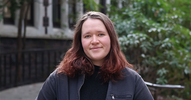 A woman is pictured in front of a stone building and ferns.