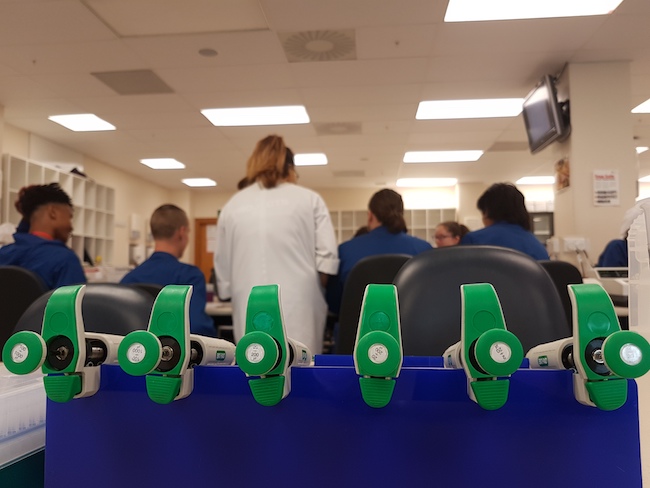 Photo of a line of six green-handled pipettes with a lab class in the background.