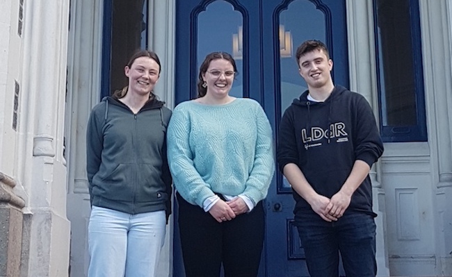 Three students standing in front of an old stone high school building.