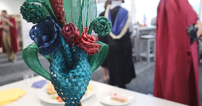 A colourful arrangement of woven harakeke items in a vase, with graduands in regalia seen in the background.
