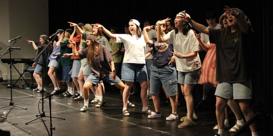 A line of young women singing and performing actions on stage while dressed in deliberately casual streetwear