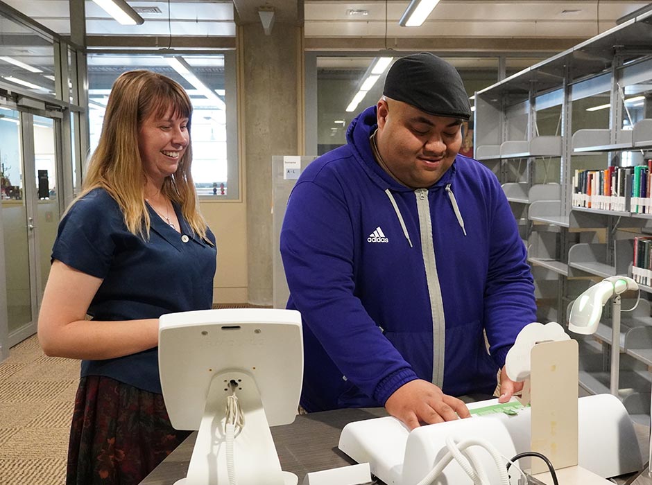 A female staff member and male student demonstrate how to check out a book at the Library.