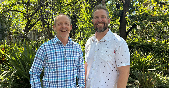 Two man are pictured outside in front of greenery.