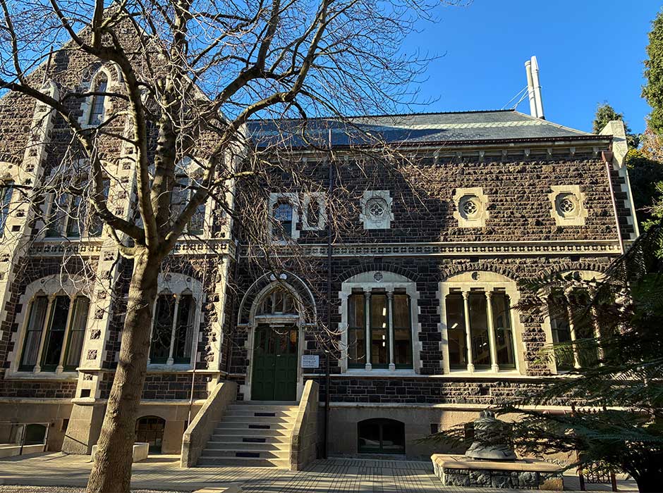 An old stone building with a sunny blue sky behind it and a couple of trees with no leaves in front of it