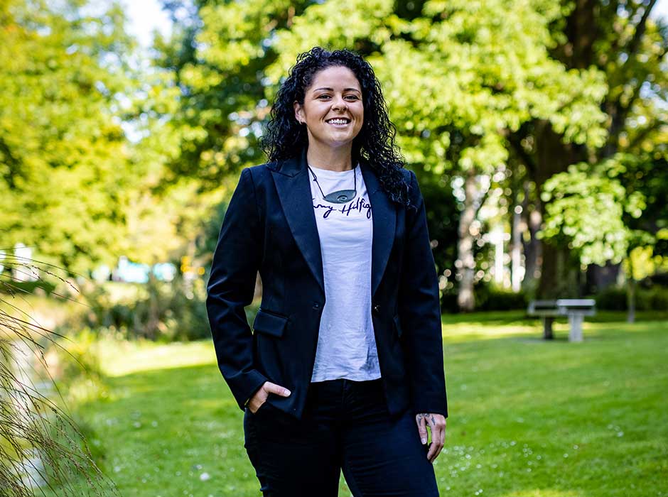 A woman standing on grass in a dark blue suit, smiling, with sunlight greenery and blue sky in the background