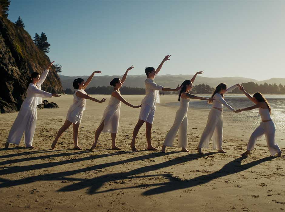 Seven dancers stand in a line on a beach 