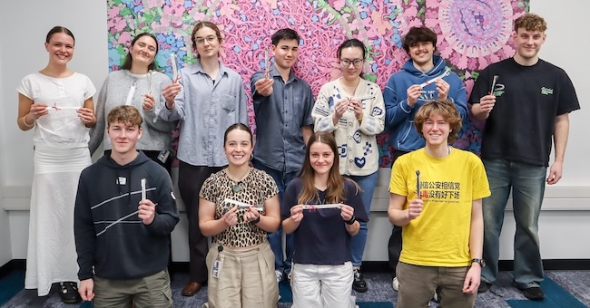 Group photo of eleven smiling students posed in front of a large, colorful scientific illustration mural