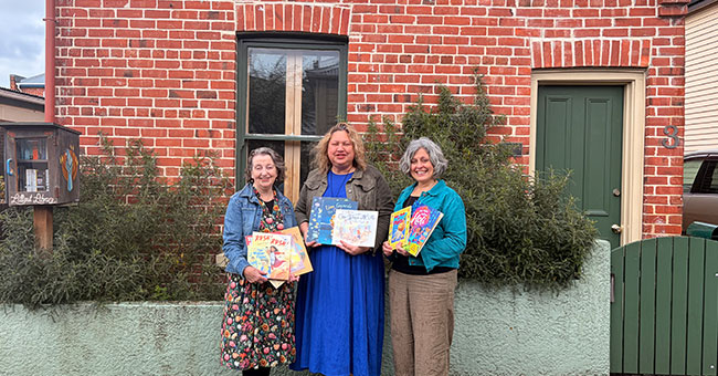 Three women hold books outside the Robert Lord Writers Cottage.