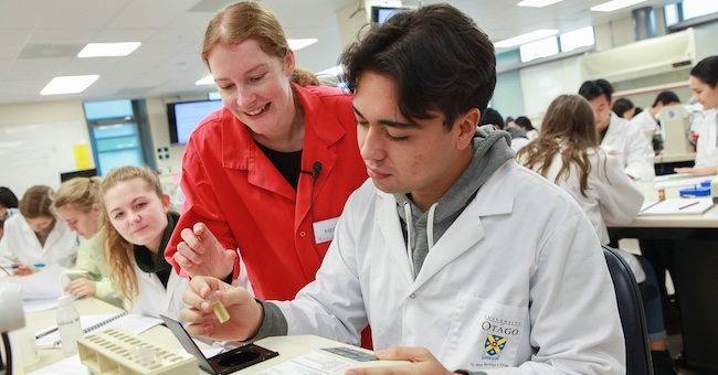 Several young people in white coats working in a science laboratory, with a teacher in a red coat offering assistance to one of them