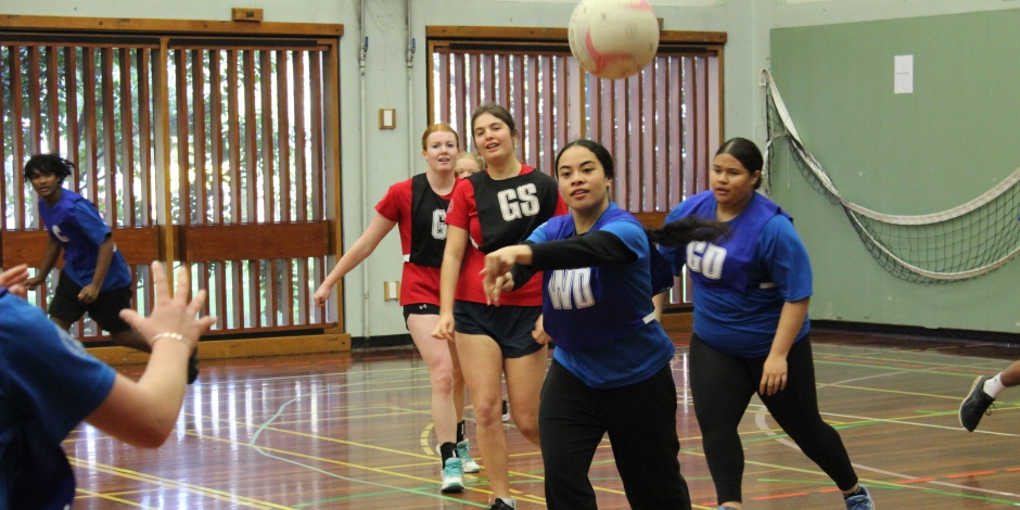 Young women in red and blue on a court. One of the blue players is passing the ball to a teammate. 