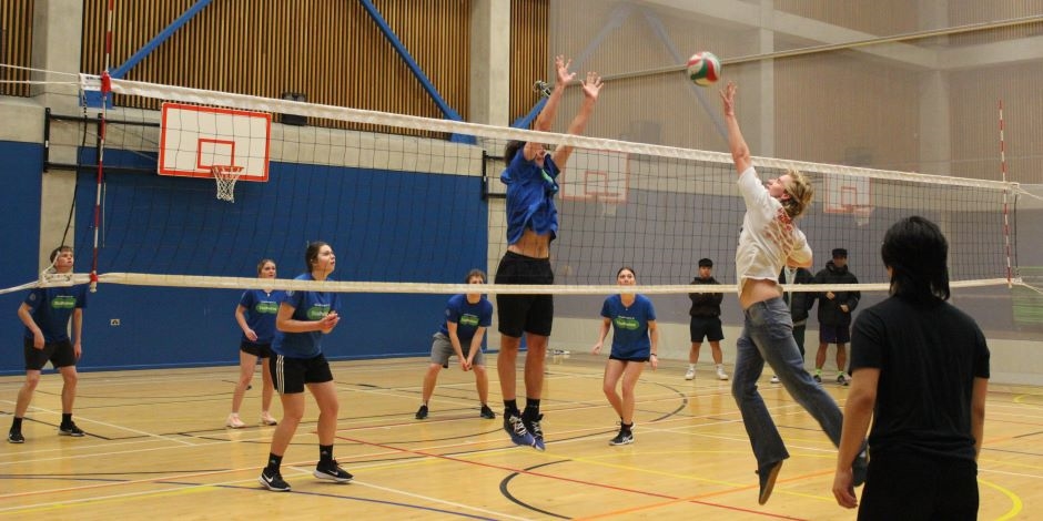 A person on each side of the net jump, one in blue to block, the other dressed in jeans and t-shirt tries to slam the volleyball. Exciting stuff.