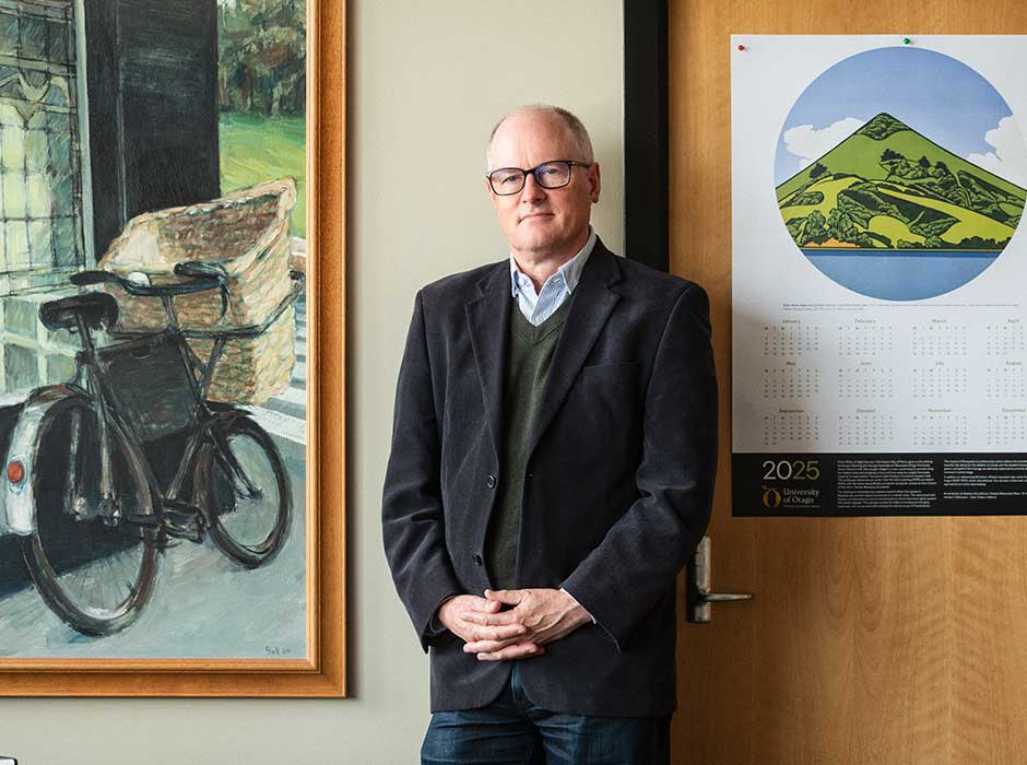 A man standing in front of a wall with a painting and a calendar behind him