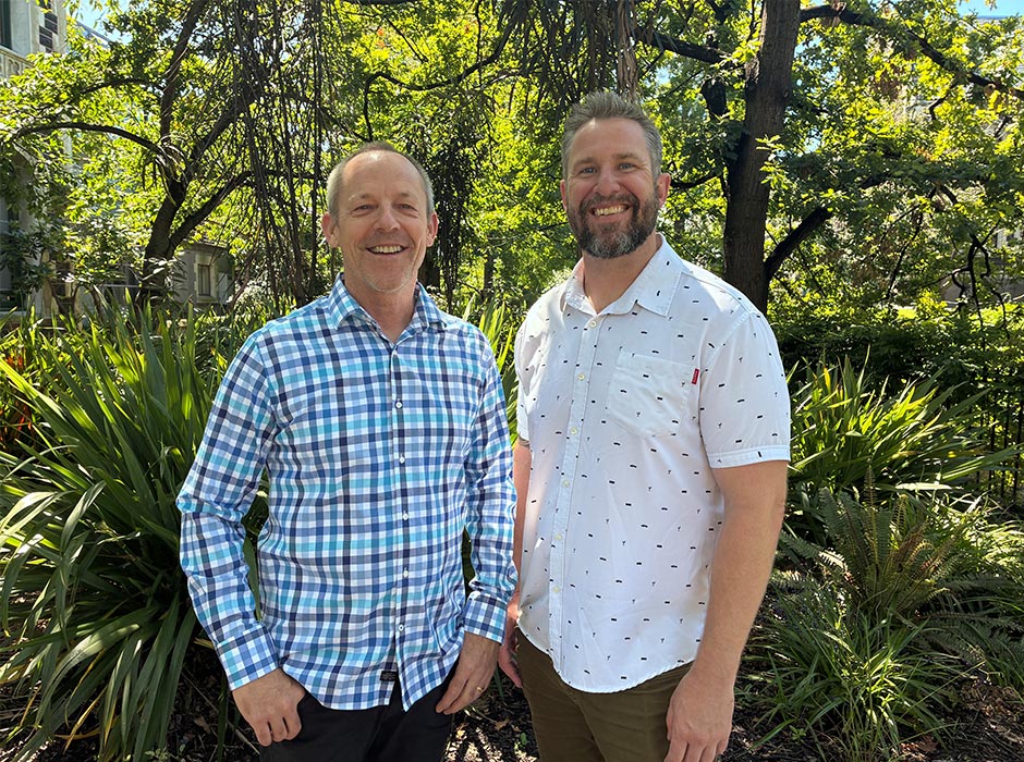 Two men are pictured outside in front of greenery.