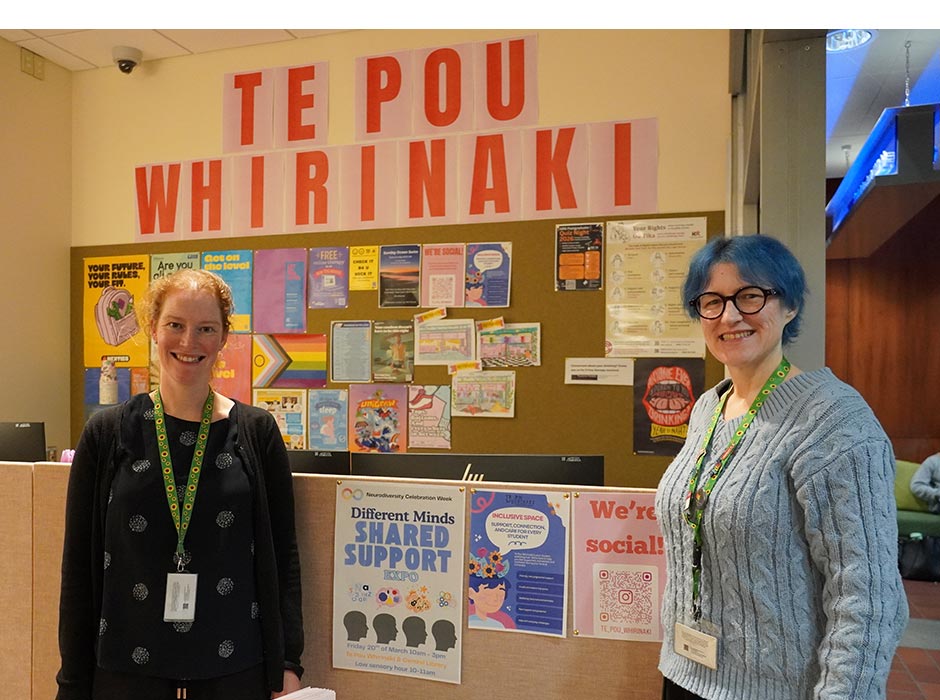 Two women stand in front of a poster display in Te Pou Whirinaki. 