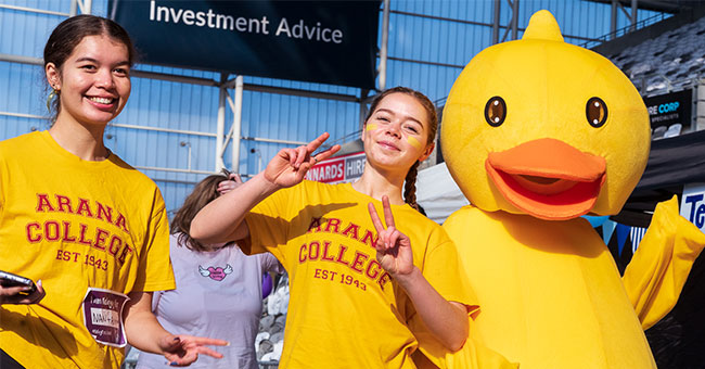 Two young women in Arana College T-shirts pose with a duck mascot.