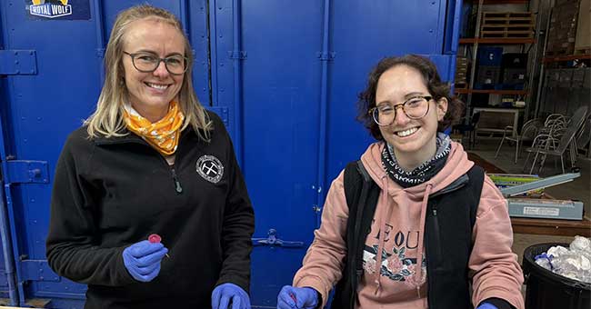 Two women standing behind a bench wearing gloves, with samples of mud on the table