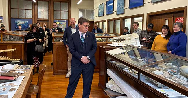 A man standing on a wooden floor in between display cases in a museum speaking to people outside the cases