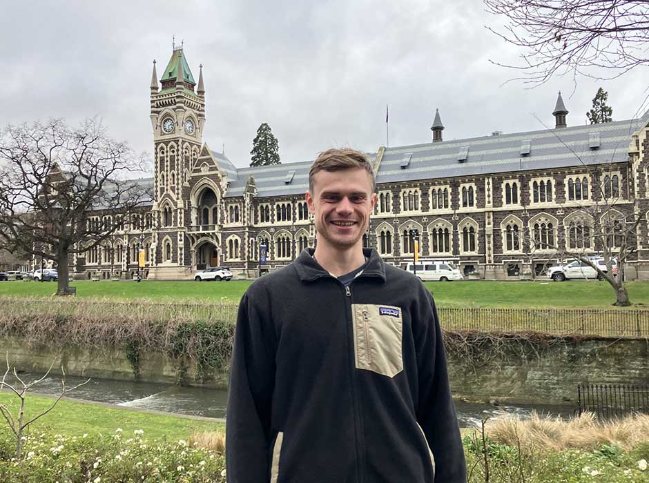 A young man standing in front of a garden bed with the University clocktower building behind him 