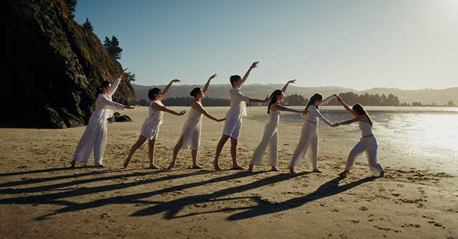 Seven dancers stand in a line on a beach 