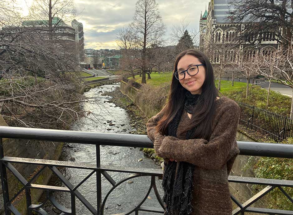 A woman stands by a handrail on a bridge, over a river with buildings in the background