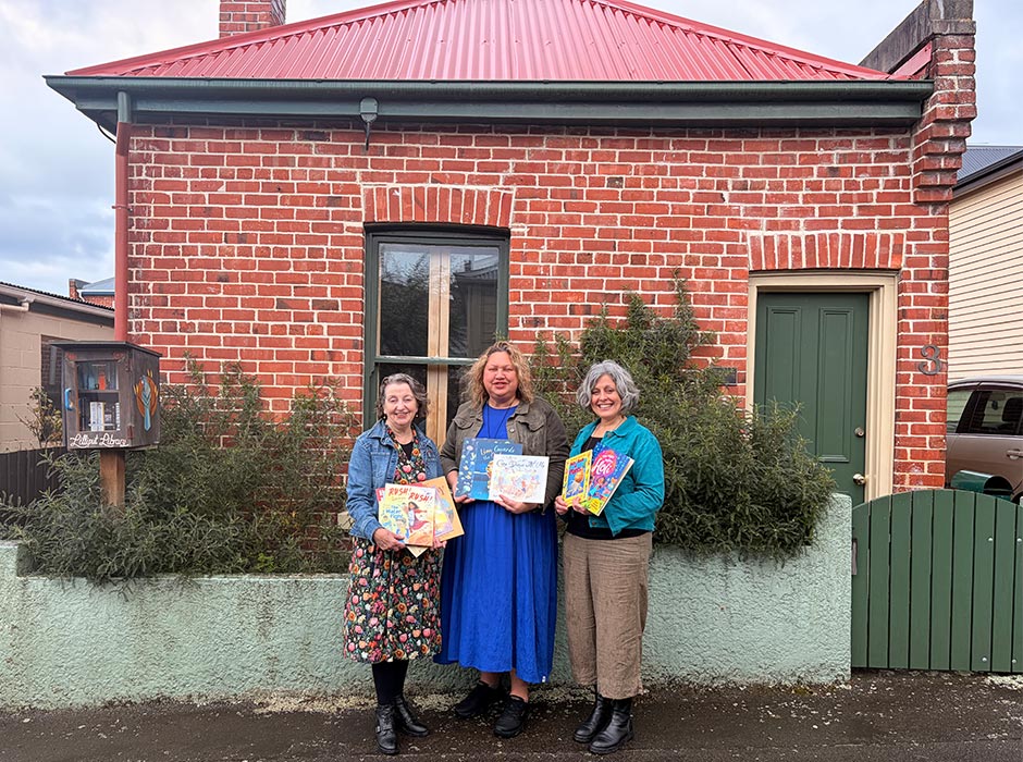 Three women hold books outside the Robert Lord Writers Cottage.