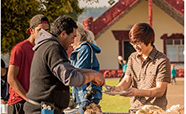 A person being served food outside with a marae in the background image