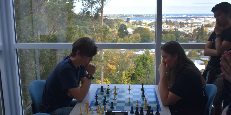Two people playing a game of chess in a residential college while another person watches.