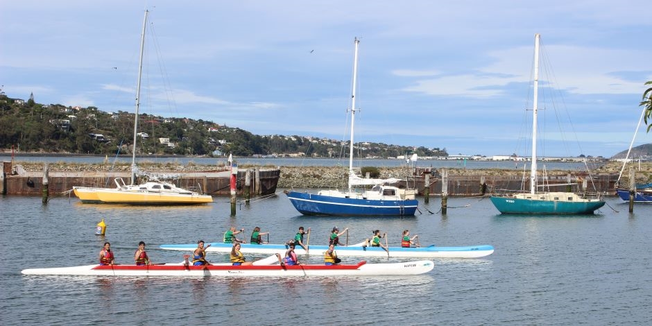 Two modern white canoes, one striped red, the other blue line up before a race in the harbour.