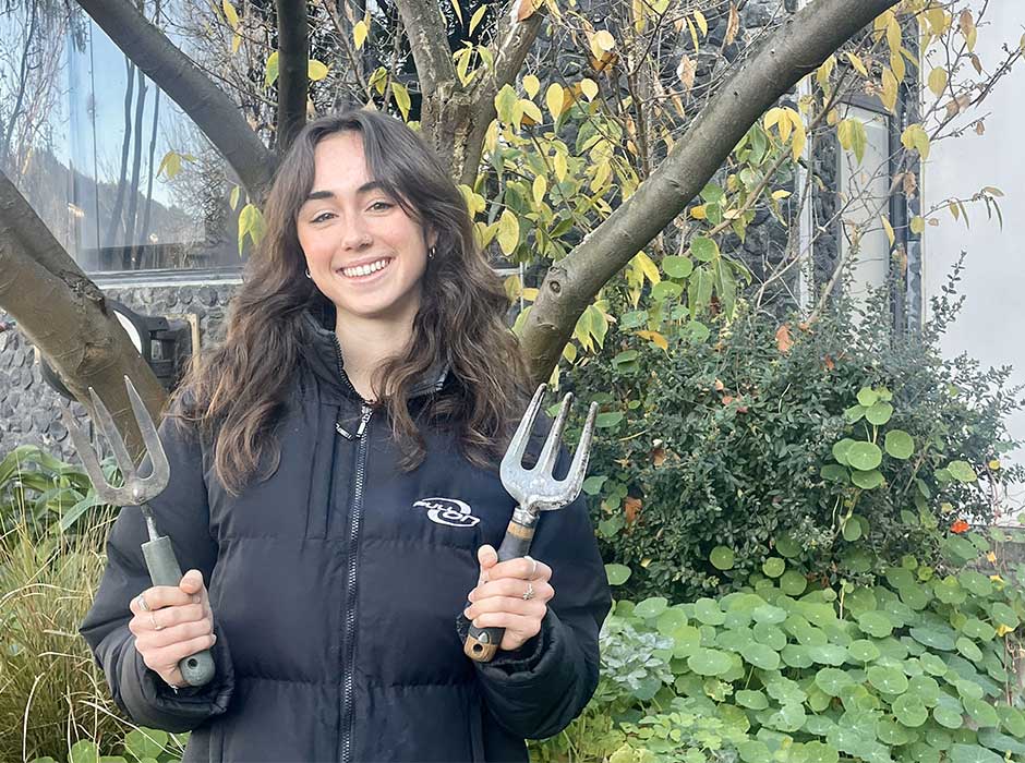 A woman standing in front of greenery holding gardening tools