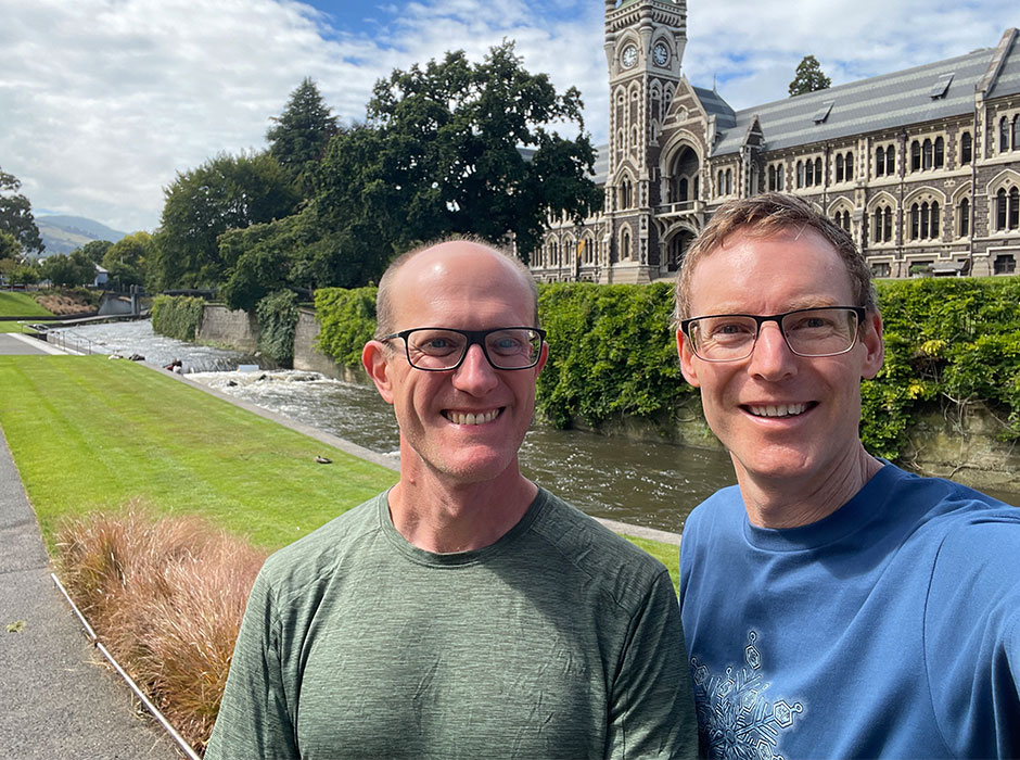 Two men standing next to each other in front of the river and clocktower