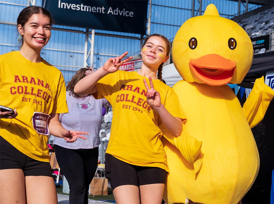 Two young women in Arana College T-shirts pose with a duck mascot.