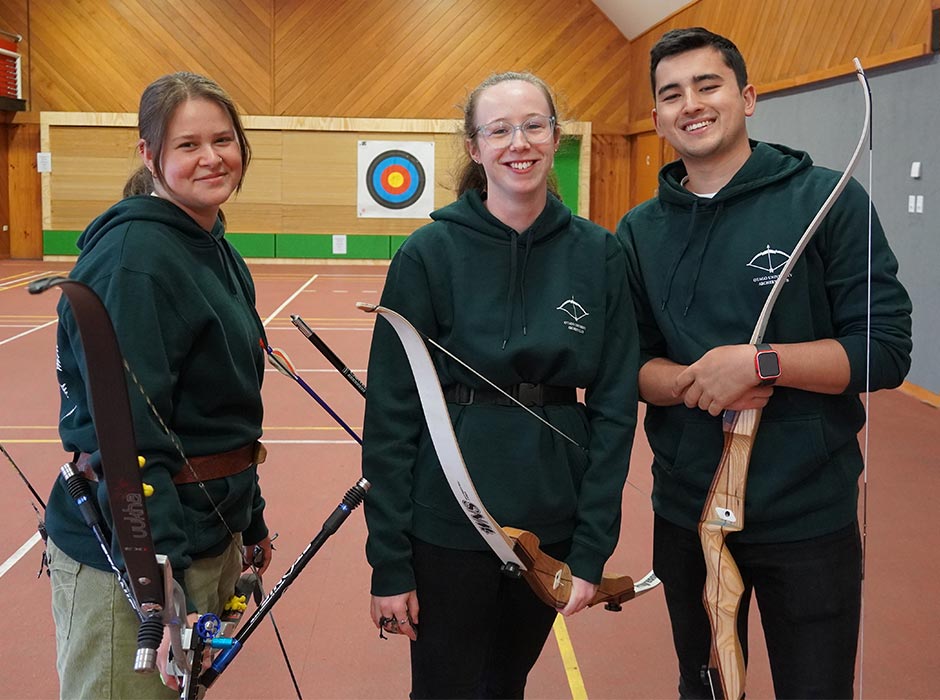 Two women and a man pose with archery equipment in front of an archery wall.