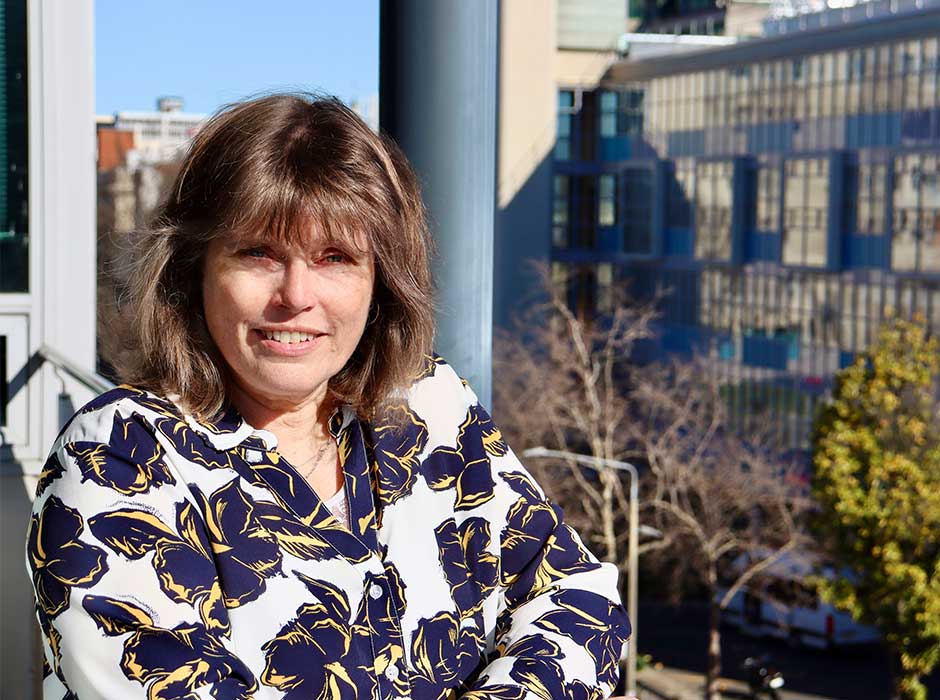 A woman from the torso up standing on a physiotherapy school building balcony on a sunny day with the dental school building in the distance