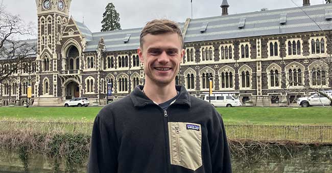 A young man standing in front of a garden bed with the University clocktower building behind him 