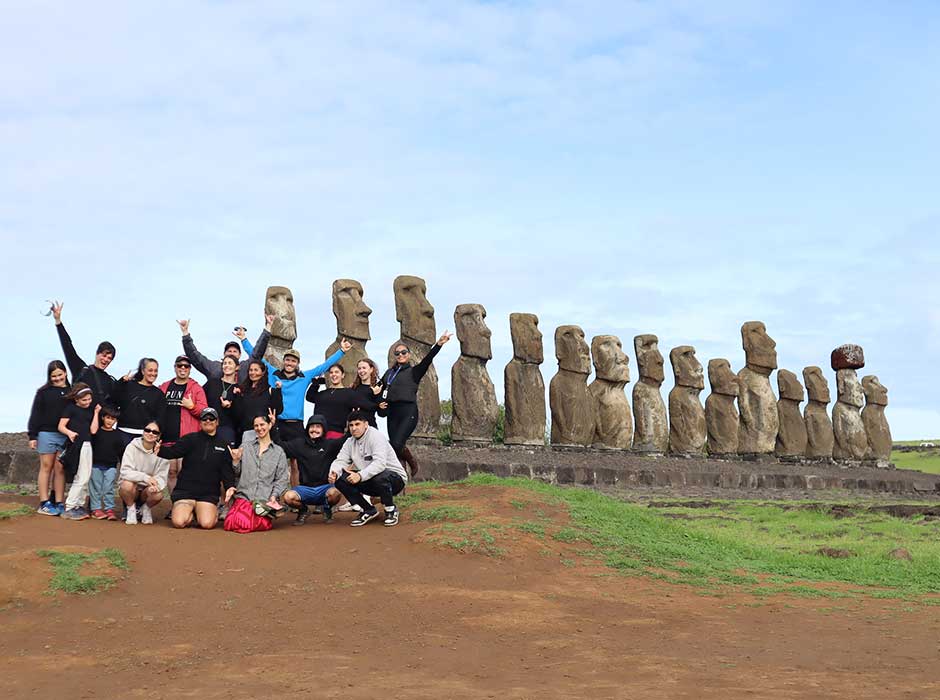 a group of people, crowded together, smiling and some waving at the camera, with a row of large stone monuments behind them