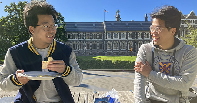 two men on a bench in front of the clocktower, one holding a sandwhich 
