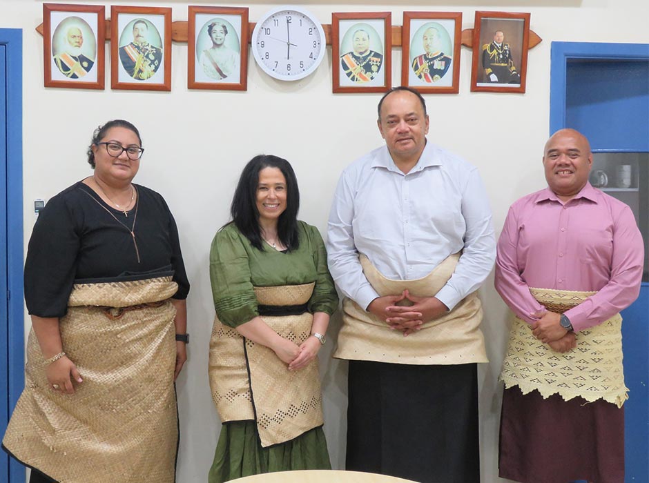 A group of people - two women and two men dressed in traditional Tongan attire, standing in front of a wall with photo frames mounted. 