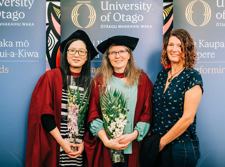 Three woman standing next to each other, two in graduation attire, in front of University of Otago banners