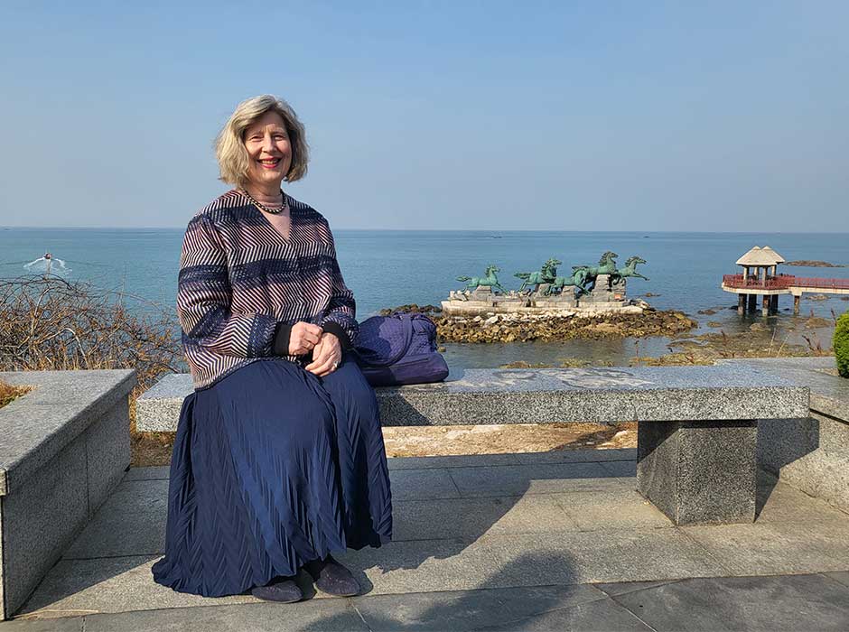 A woman seated on a concrete bench in front of the ocean on a nice sunny day, there are statues of horses, made of oxidised copper, on a small island in the background