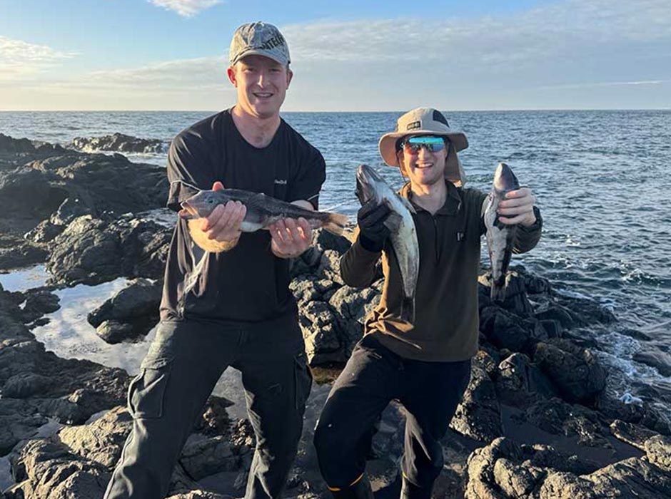 Two men, standing on a rocky outcrop holding fish, with the sea behind them.